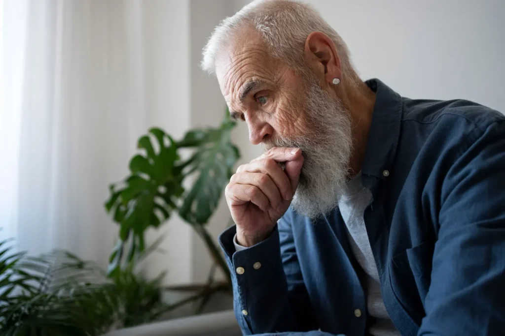 Older man wearing a navy blue outfit, sitting with a thoughtful or worried expression, reflecting concerns about aging and health, particularly prostate or urinary issues.
